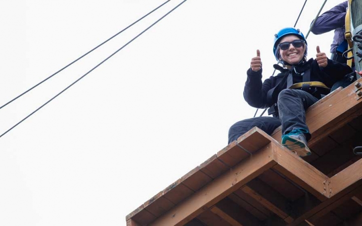 A person wearing safety gear and secured by ropes sits atop a wooden structure of a high ropes course, looks down and gives the camera two thumbs up.